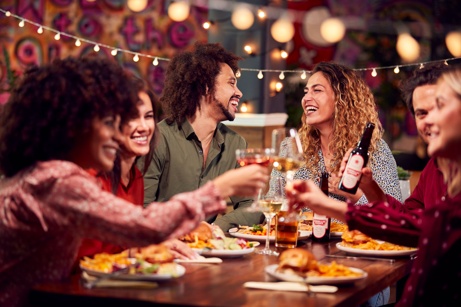 Group of friends celebrating with drinks and food at an outdoor restaurant with string lights